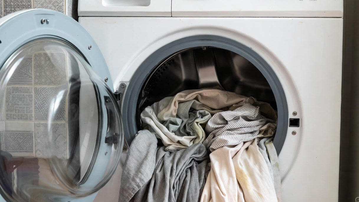 Landscape image showing a household washing machine with delicate home fabrics like curtains and cushions visibly creased and distorted, illustrating why home washing instead of professional dry cleaning can be risky for non-clothing items.
